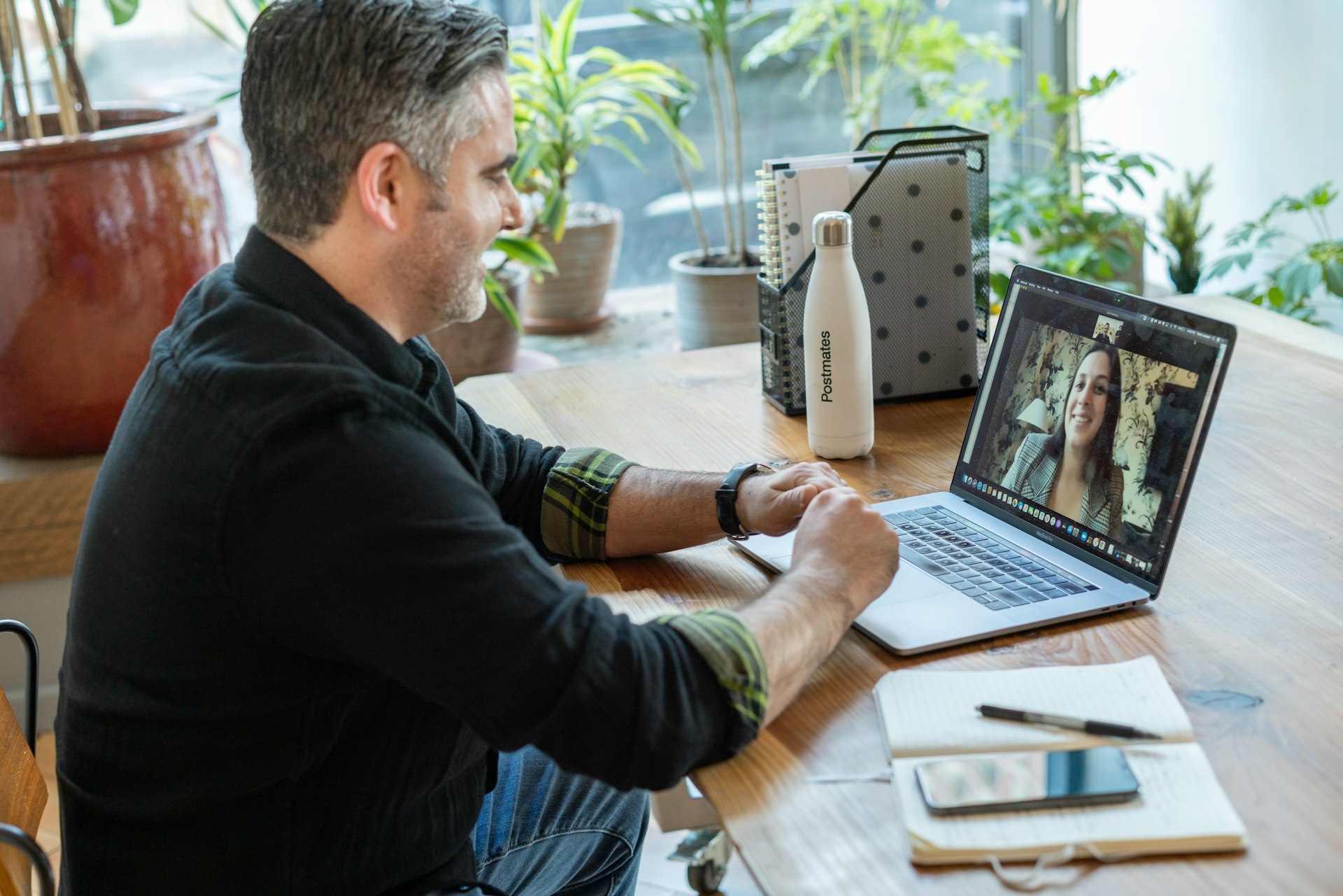 Man participating in a virtual meeting on his laptop in a workspace with natural light and plants, taking notes.
